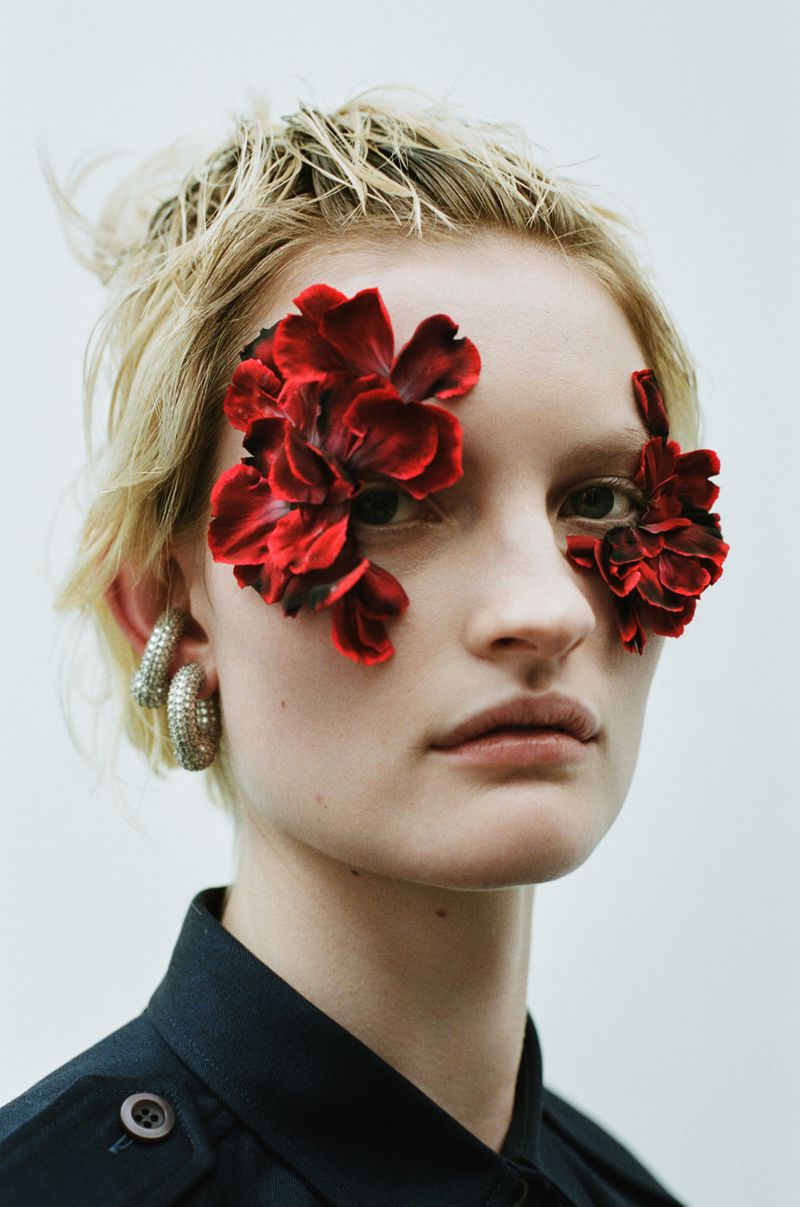 Close-up portrait of a fair-skinned woman with flowers covering her eyes, wearing hoop earrings and a black collared shirt indoors.