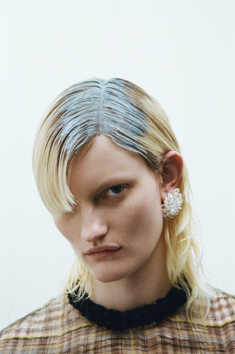 Close-up portrait of a blonde-haired woman with a geometric hair dye pattern and floral earrings against a stark white backdrop.
