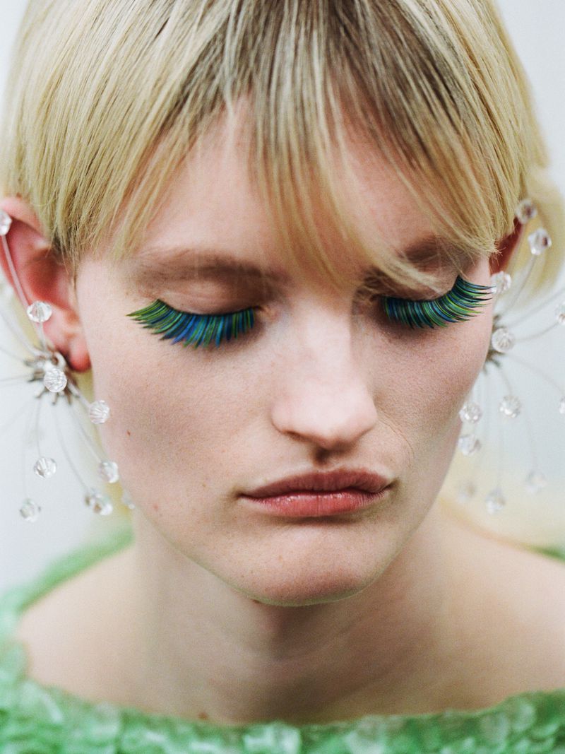 Close-up portrait of a fair-skinned adult female with creative green and blue eyelashes, light blonde hair, and decorative earrings.
