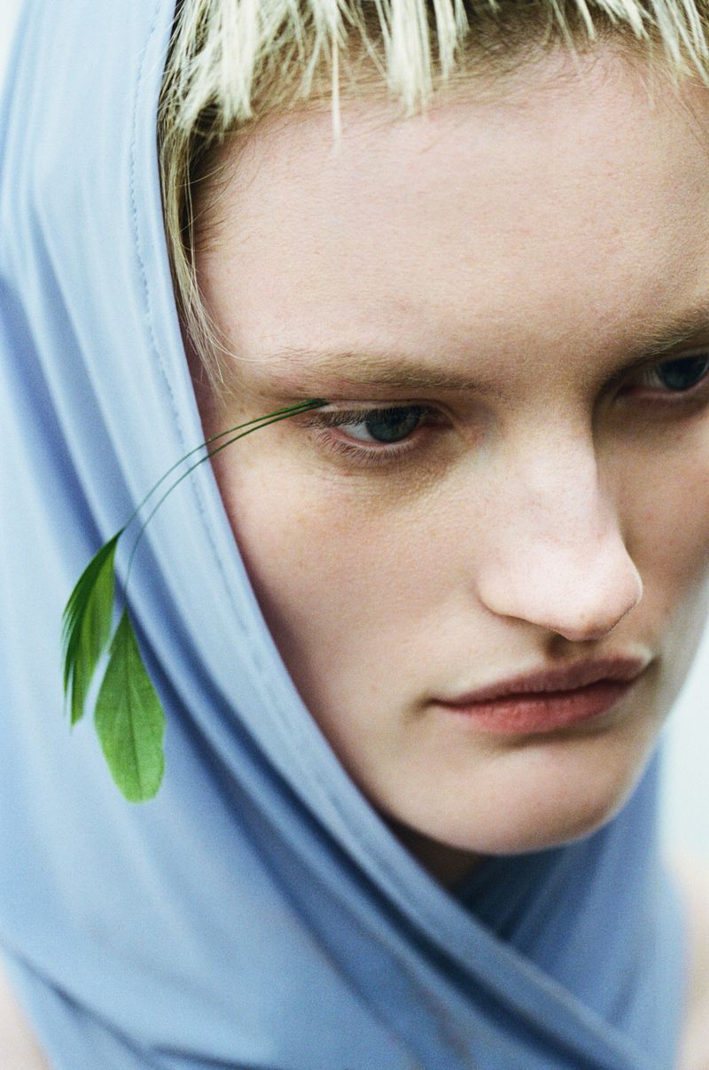 Close-up of a woman with light skin and blonde hair wearing a light blue head covering and a green feather decoration near her eye.