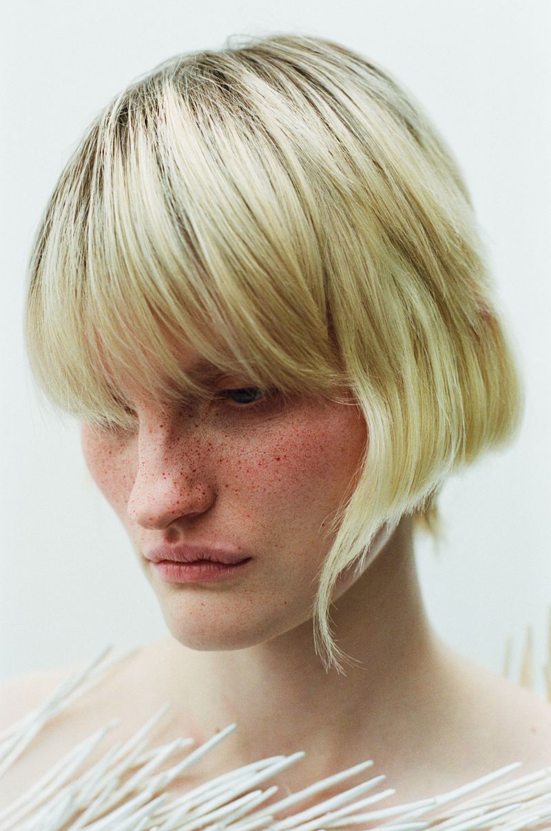 Close-up portrait of a fair-skinned adult female with blonde bob haircut, freckles, and white fibers in front of a neutral background.
