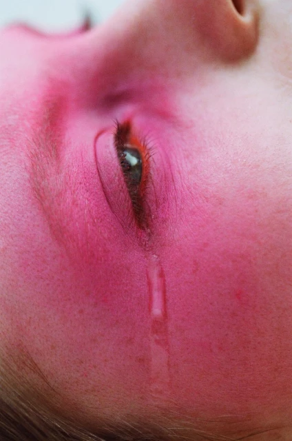 Close-up portrait of a weeping adult female with red-toned skin, one eye open with a tear rolling down her cheek.