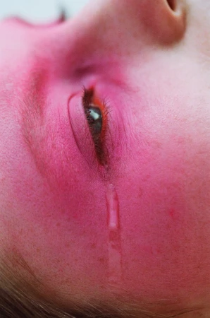 Close-up portrait of a weeping adult female with red-toned skin, one eye open with a tear rolling down her cheek.