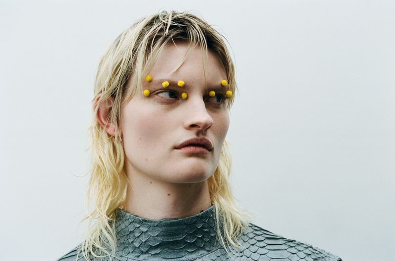 Close-up portrait of a blond adult female with yellow decoration above her eyebrows, wearing a grey snakeskin patterned top.