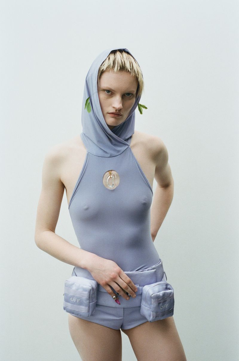 Fashion portrait of a slender female adult model in a blue bodysuit and head covering against a light background in a studio.