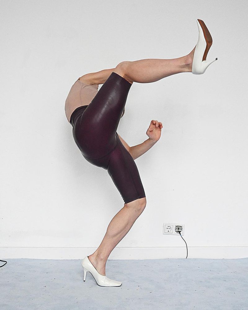 Adult wearing latex shorts and white high heels is in a bent over balancing pose against a white backdrop inside an empty studio.