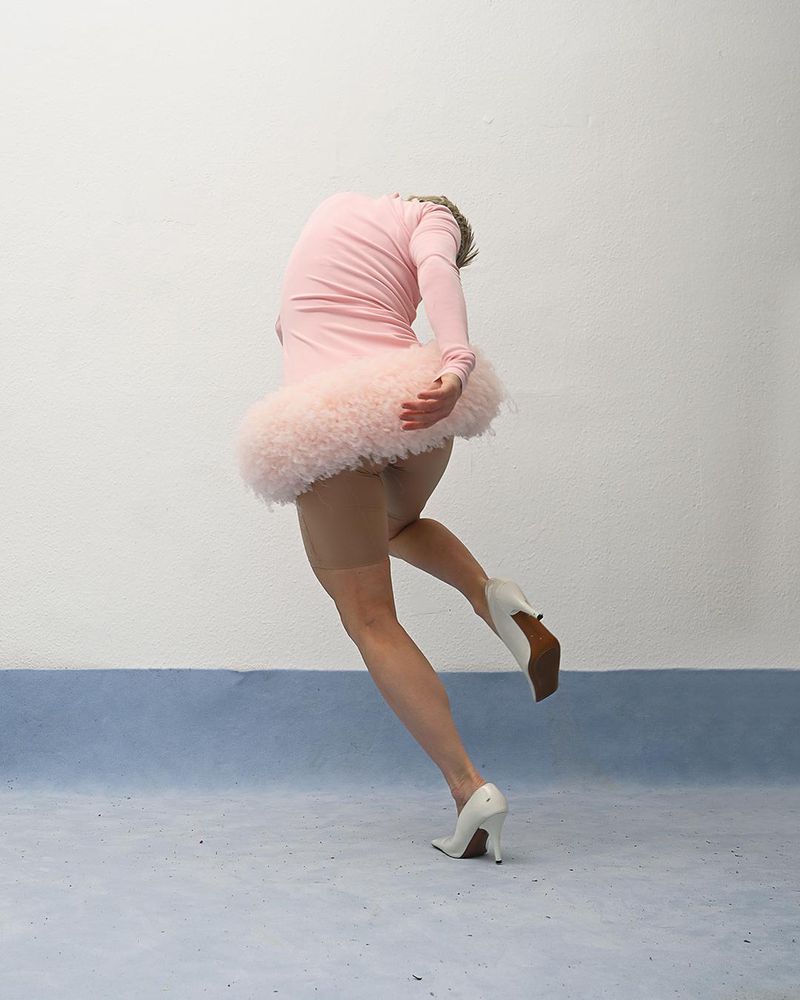 Model in motion wearing a pink long-sleeve leotard with a fluffy trim and white heels, against a two-tone white and blue wall.