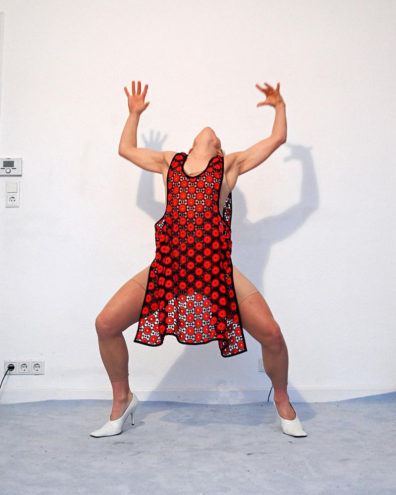 Person in squat pose wearing red crochet dress and white heels in a white studio setting with arms raised overhead.