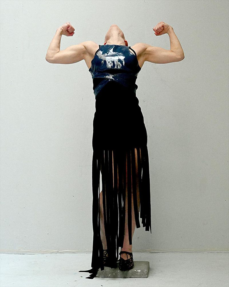 Person wearing a dark top and fringed skirt stands on a block with arms raised and head tilted back against a light wall indoors.