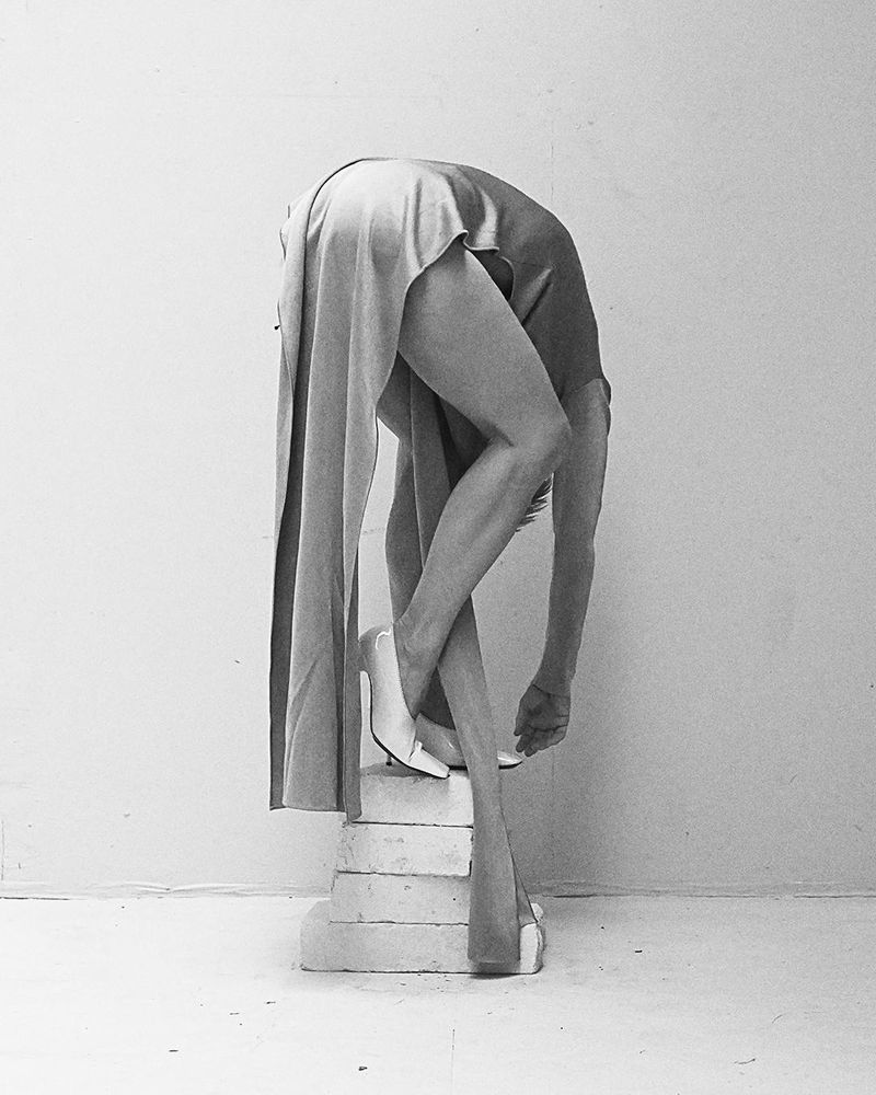 Woman in dress and heels bending over a tiered pedestal, black and white fine art fashion photography indoors with minimalist composition.