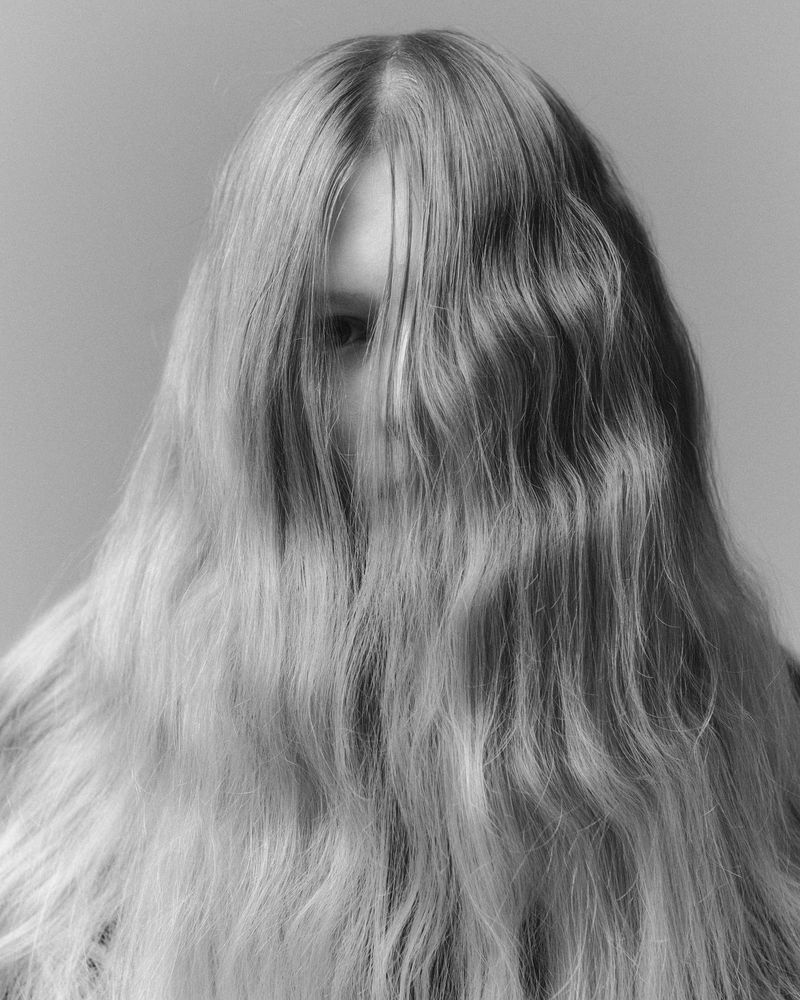 Black and white portrait of a person with long blond hair covering their face, set against a plain grey backdrop.