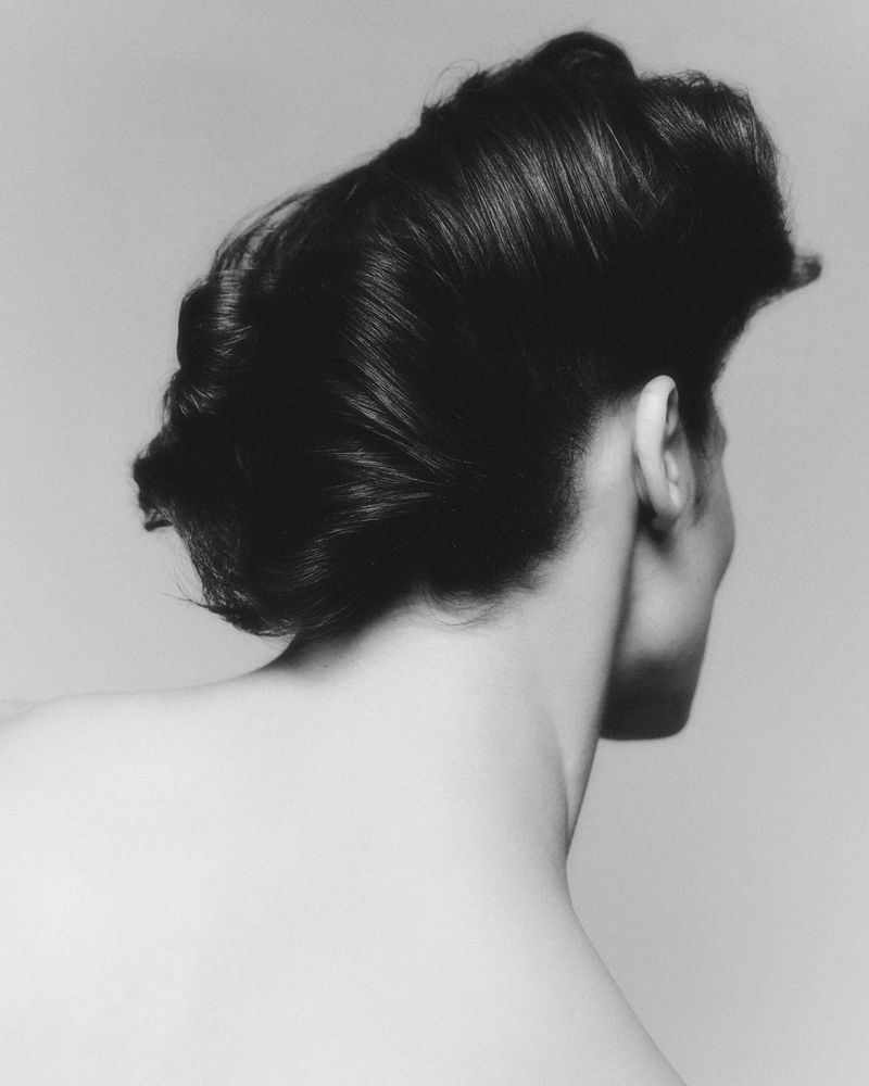 Monochromatic portrait of a person with dark, styled hair, captured from behind against a simple light background in a minimalist studio setting.