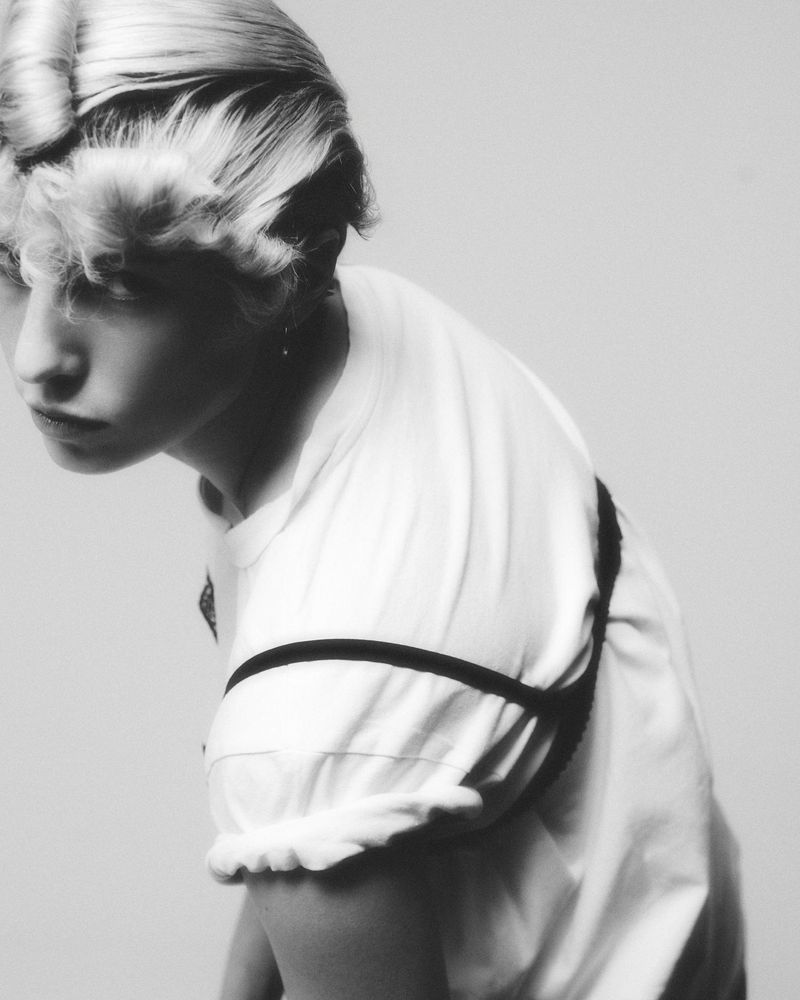Black and white portrait of a young adult woman with styled blonde hair wearing a white t-shirt and dark shoulder straps in a studio setting.