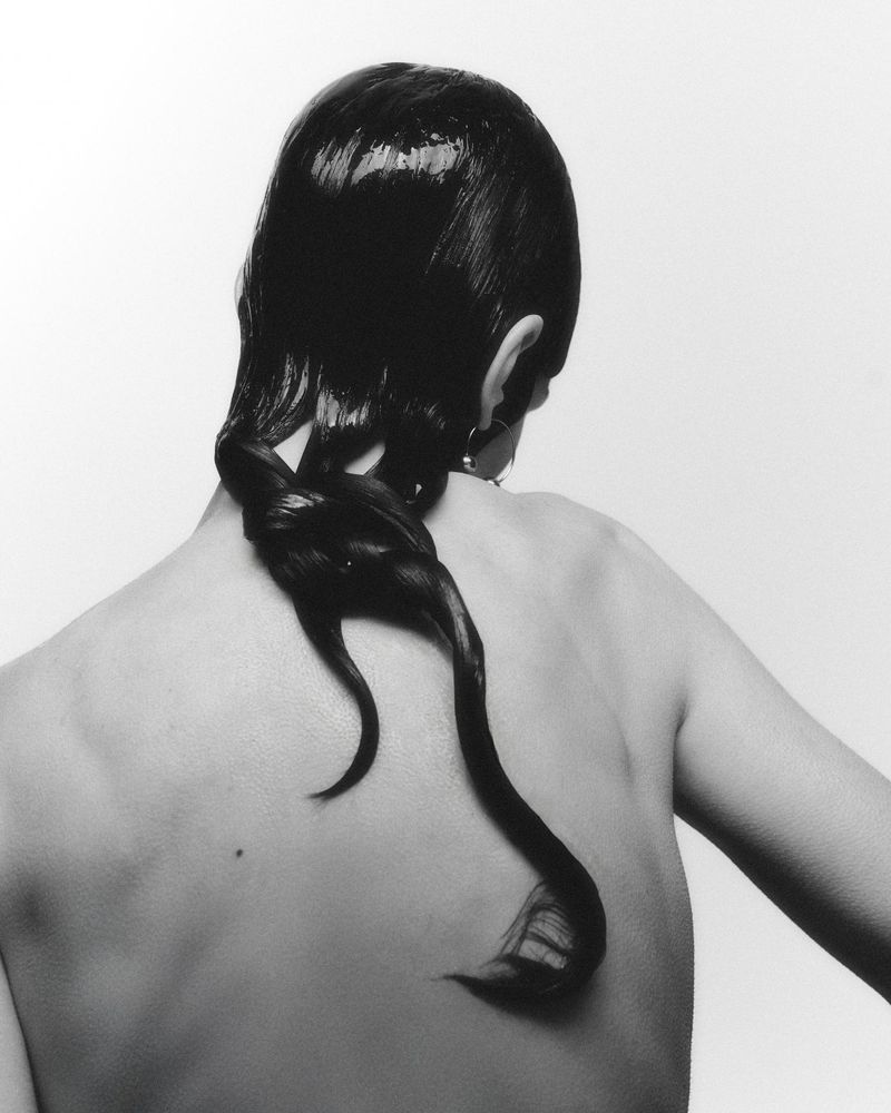 Back view of an adult woman with styled wet-look black hair twisted into a partial ponytail, posing in front of a white background.