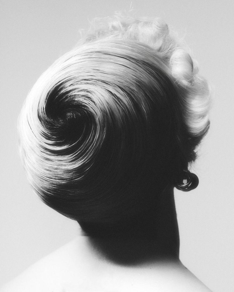 Black and white studio portrait from behind showing a sculptural spiral chignon hairstyle on a blonde woman with an earring.
