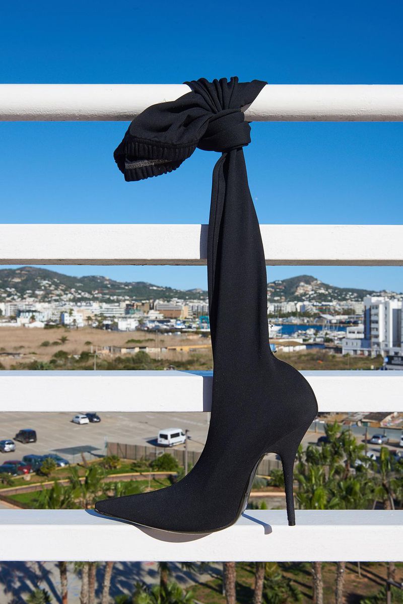 Black high heel sock boot and knotted hat on a white railing with a sunny cityscape and blue sky in the background.