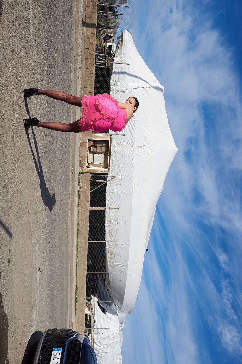 Woman in a bright pink dress and black tights poses on a street next to a large white tent on a sunny day with a blue sky.