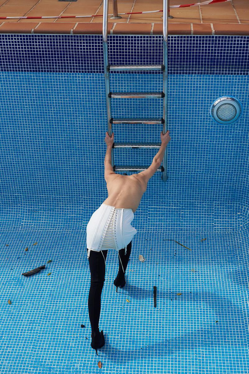 A woman in a white corset and heels climbs a ladder in an empty, blue-tiled swimming pool, creating a surreal and fashion-forward image.