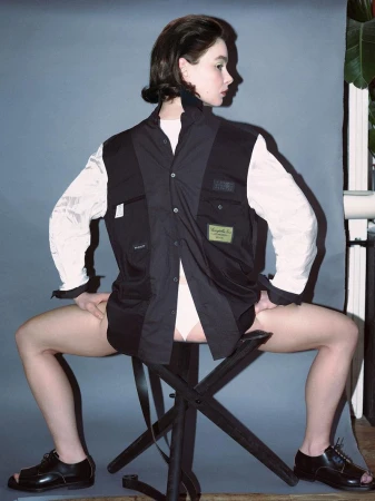 A stylish young woman in a black and white shirt sits on a black folding stool in a studio against a grey background, wearing black shoes.