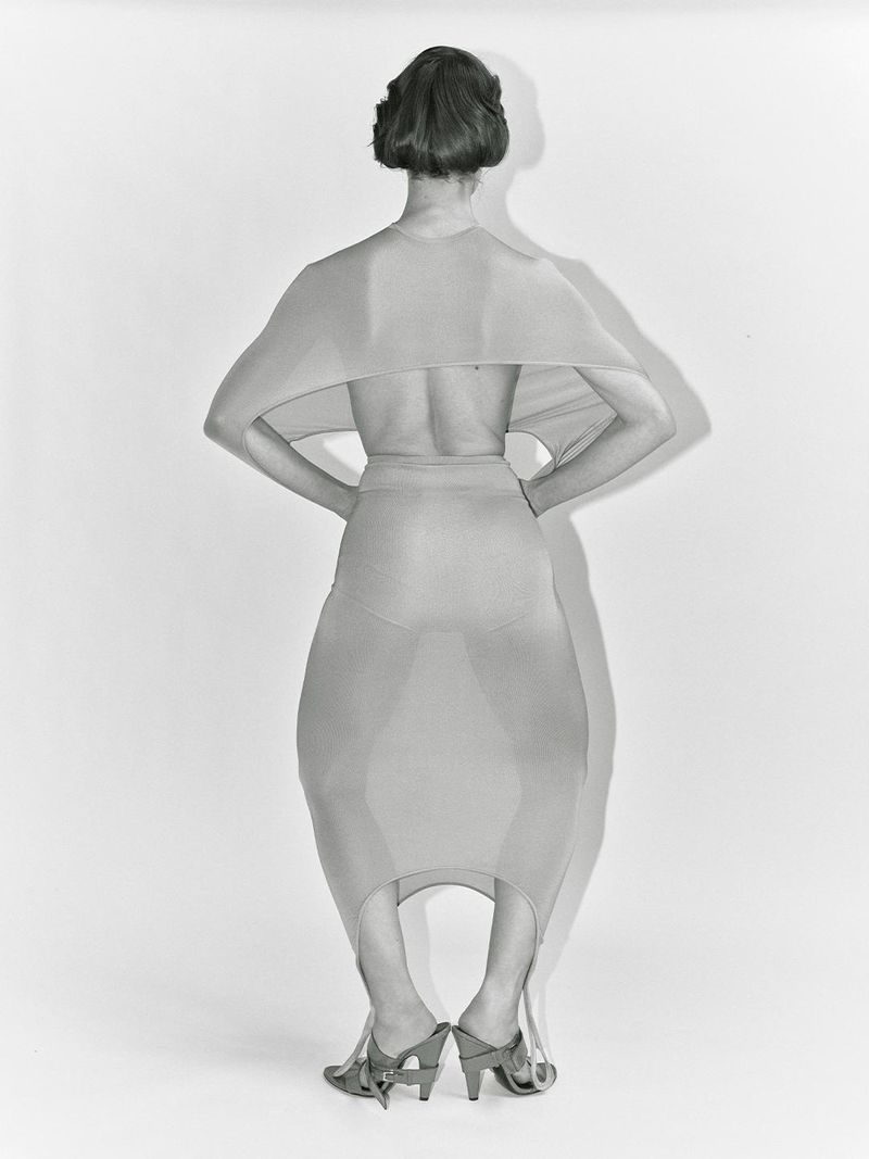 Back view of a woman in a beige dress and heels, hands on hips, standing in a studio with a neutral backdrop, with shadows.