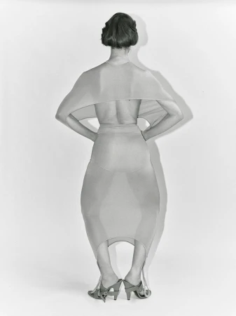 Back view of a woman in a beige dress and heels, hands on hips, standing in a studio with a neutral backdrop, with shadows.