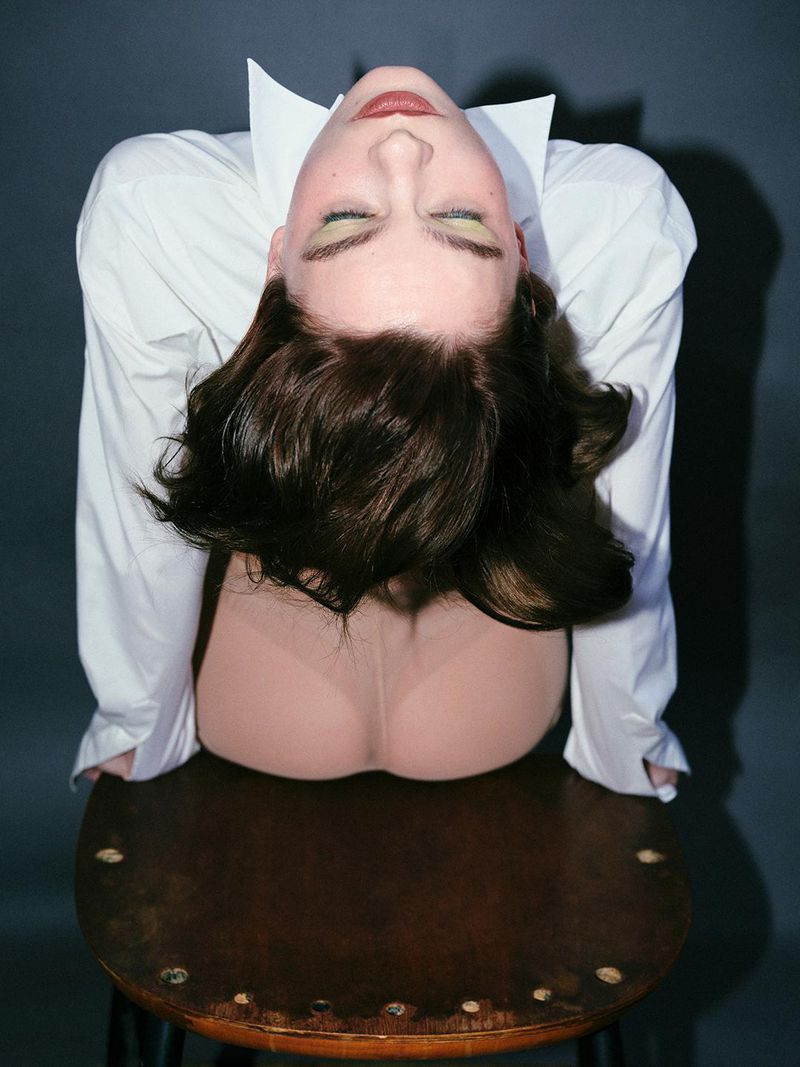 Adult woman with brown hair poses sitting on a stool, eyes closed, wearing a white shirt, in a studio with a grey background.