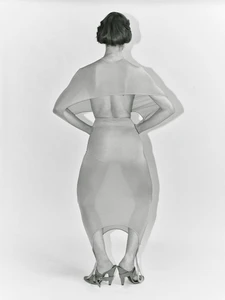 Model seen from the back wearing a sheer sculptural draped outfit and high heels against a white studio background.