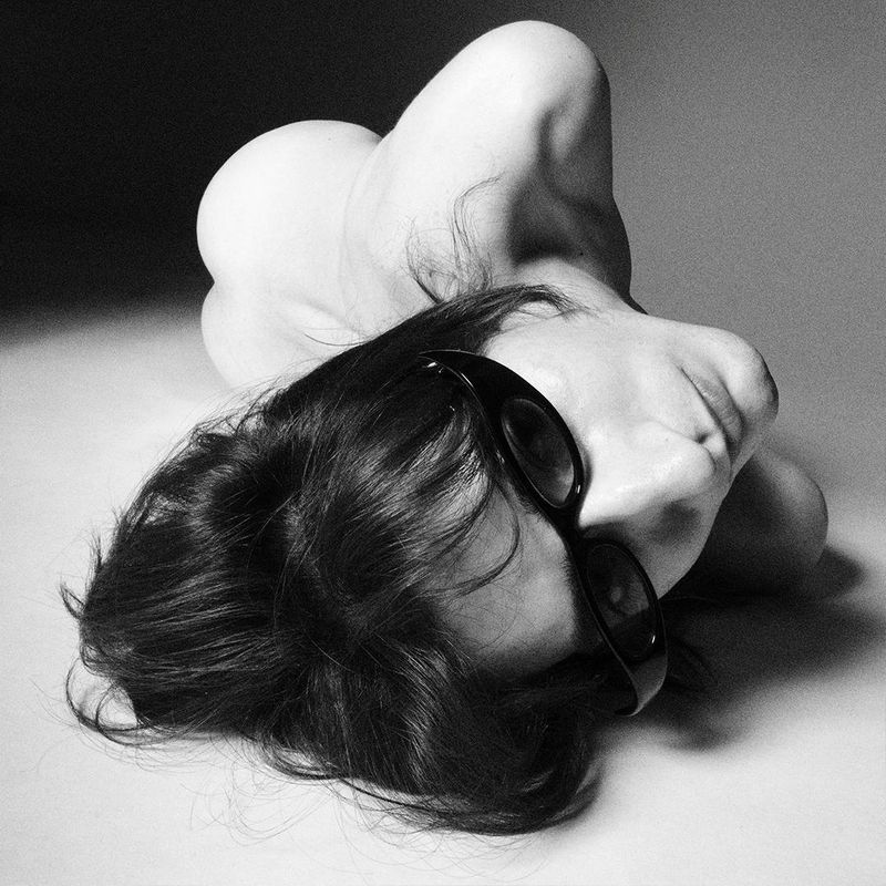 Black and white portrait of an adult laying down wearing sunglasses. Body is contorted and folded into itself, against a dark background.