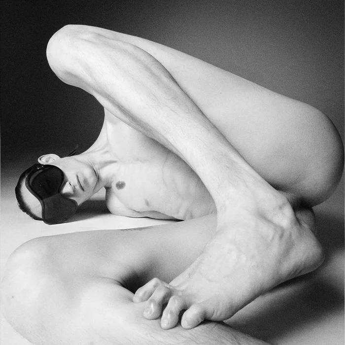 Sculptural black and white portrait of a contorted nude figure wearing a black eye mask against a neutral studio backdrop.