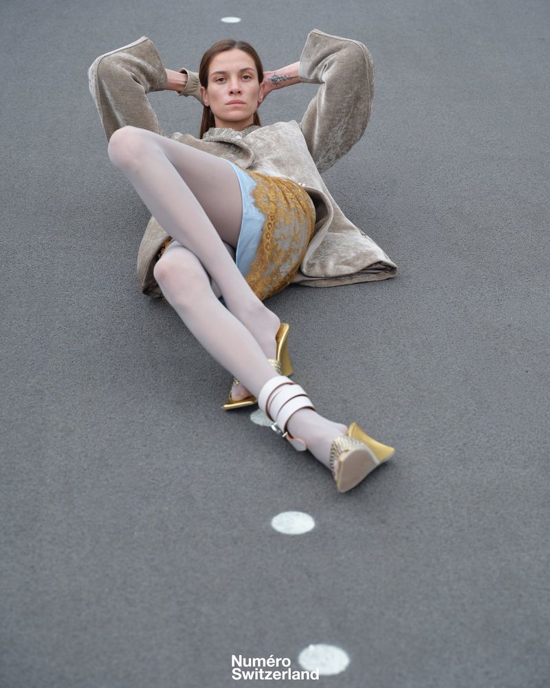 Fashion editorial shot of a woman in hosiery and jacket reclining on a speckled gray surface in gold heels with white straps. 