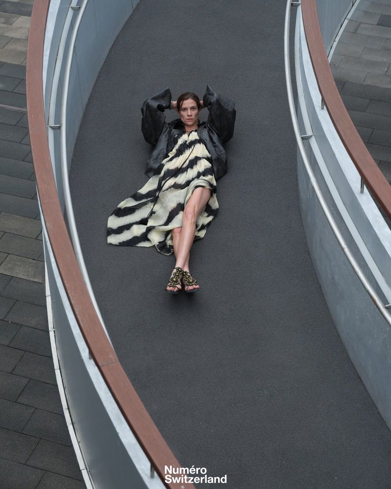 A high-angle shot showcases a model reclining on a curved walkway, wearing a zebra-print dress and black jacket for a fashion editorial.