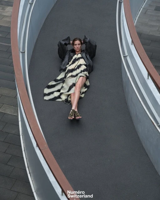 A high-angle shot showcases a model reclining on a curved walkway, wearing a zebra-print dress and black jacket for a fashion editorial.