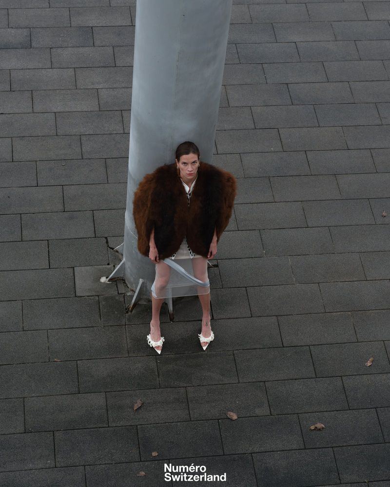 Eye-level view of a young woman in a fur coat and heels posing by a gray column on a paved surface for a fashion editorial.