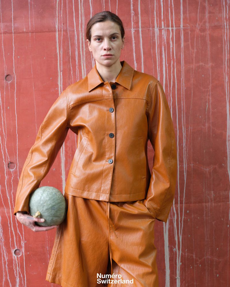 A poised woman in a tan jacket and skirt holds a melon against a concrete wall, showcasing editorial fashion in natural lighting.