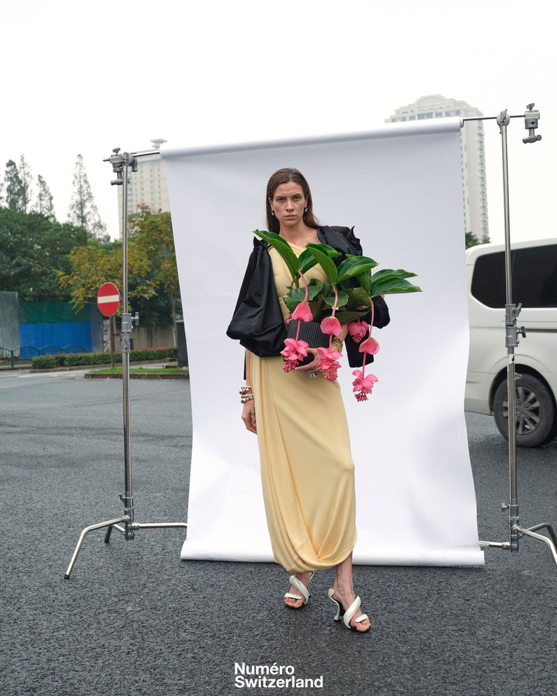 Adult female in dress and jacket holding a pink flowering plant standing in front of a portable white backdrop outdoors.