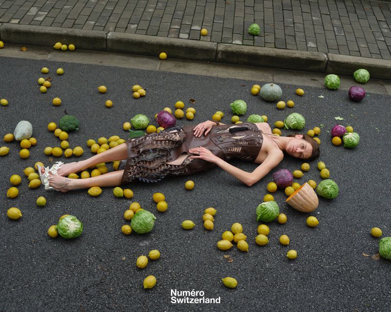 Woman in a brown dress lies on a grey asphalt surface surrounded by lemons, cabbages, and other vegetables with a brick wall background.