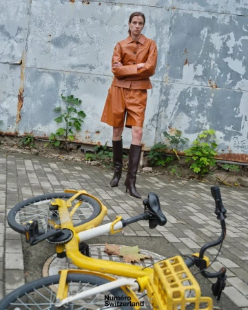 A woman in a tan leather jacket and shorts poses near a fallen yellow bike against a weathered wall in an urban outdoor fashion shot.