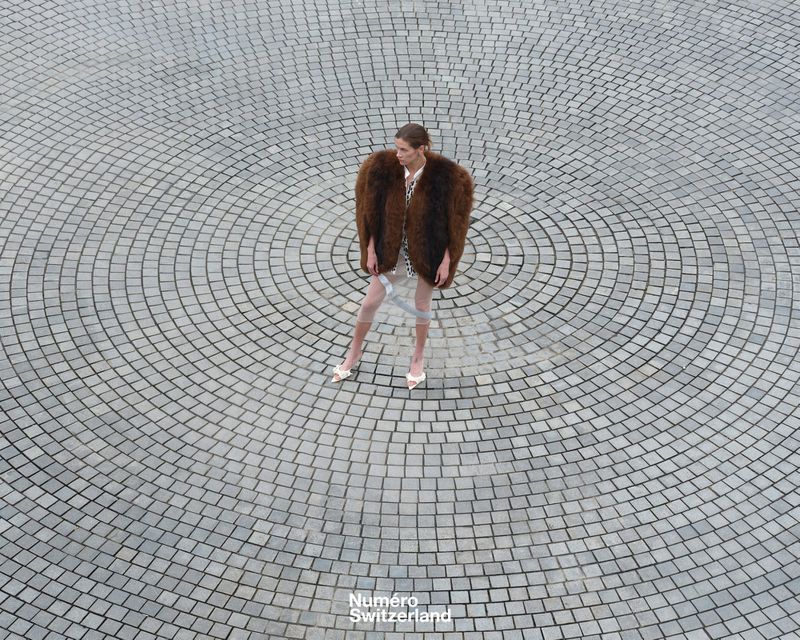 Overhead shot of a woman in a fur coat and heels standing on a circular stone-tile surface in an outdoor environment on a cloudy day.