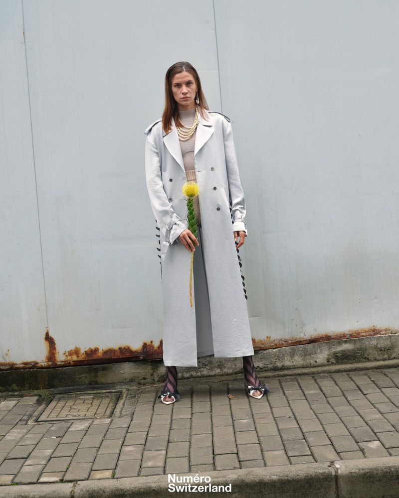A female model stands outside in a silver trench coat and patterned tights, holding a yellow flower in front of a grey background.