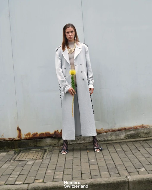 A female model stands outside in a silver trench coat and patterned tights, holding a yellow flower in front of a grey background.