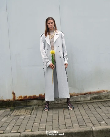 A female model stands outside in a silver trench coat and patterned tights, holding a yellow flower in front of a grey background.