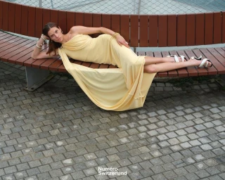 Model Vaiora Stroganoff reclining in a draped pale yellow gown on a curved wooden bench for Numéro Switzerland.