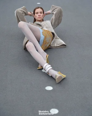 Model Vaiora Stroganoff lying on an asphalt road in a textured taupe coat, yellow skirt, and gold heels for Numéro Switzerland.