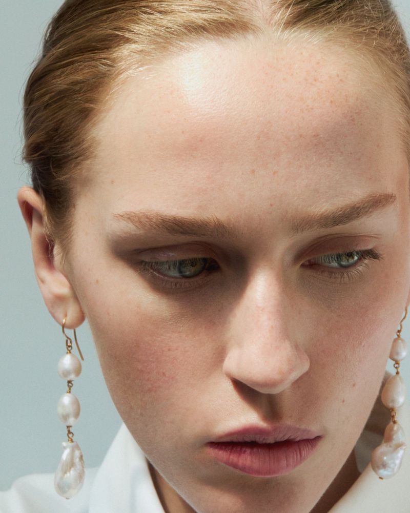 Close-up of a fair-skinned woman with pearl earrings and a downward gaze in a portrait-style image. The focus is on her facial features.