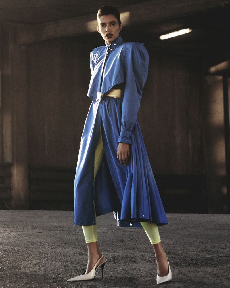 Fashion shot of a woman wearing a blue trench coat with a gold belt, lime green leggings, and white slingback heels in an urban setting.