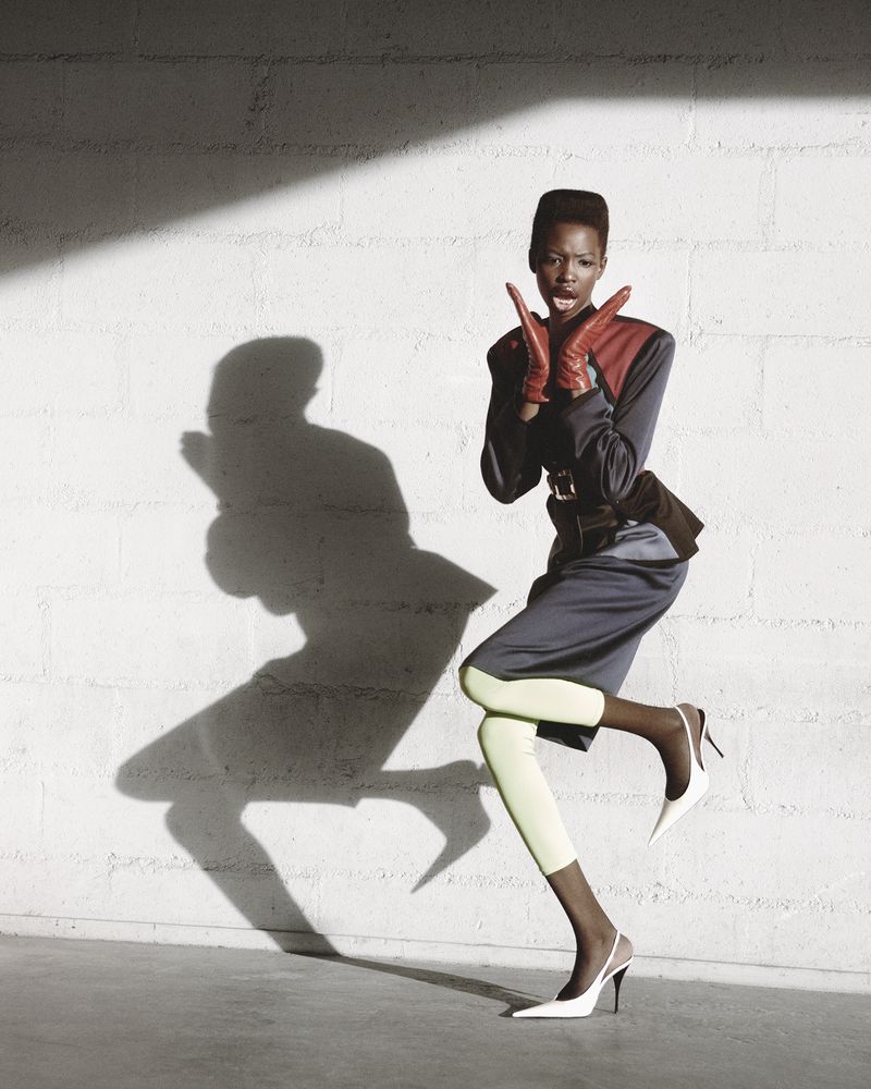 A stylish Black woman in vintage fashion poses against a white textured wall, casting a shadow, wearing gloves, skirt, and high heels.