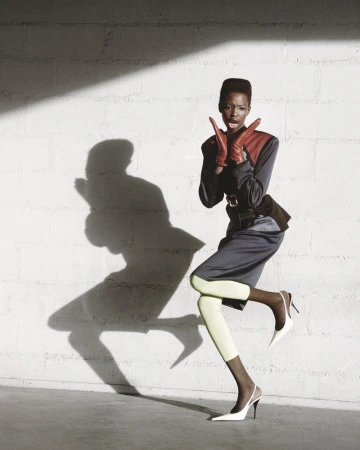 A stylish Black woman in vintage fashion poses against a white textured wall, casting a shadow, wearing gloves, skirt, and high heels.