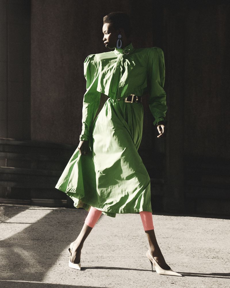 Fashion editorial shot of a Black woman in a green dress and white heels walking against a dark stone wall with shadows.