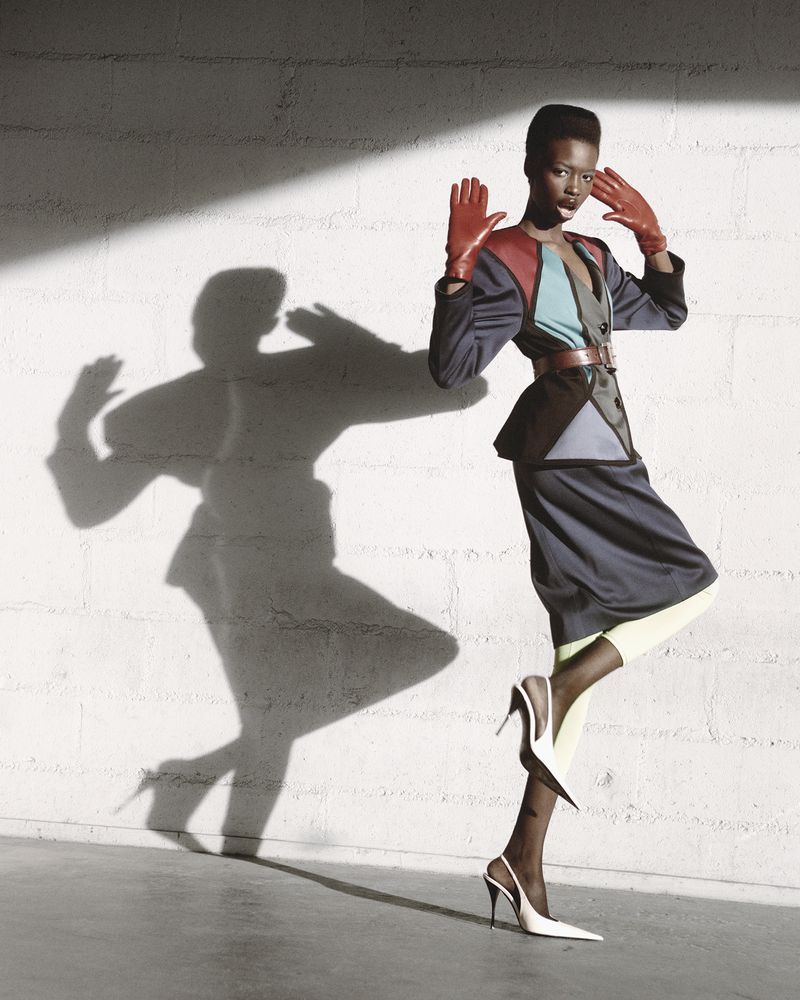 Grace Jones poses in a color-blocked outfit with red gloves, styled with white heels, in a studio setting with shadow play.