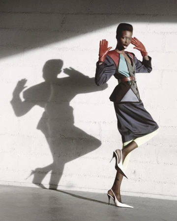 Grace Jones poses in a color-blocked outfit with red gloves, styled with white heels, in a studio setting with shadow play.